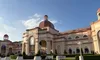 The image shows a large, elegant building with domed roofs and arched windows, surrounded by landscaped greenery and featuring a bustling entrance.