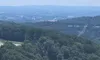 The image shows a hilly landscape with a winding road, dense green forests, power lines, and a billboard, with a hazy view of a distant city in the background.