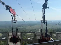 The image shows a zipline platform with harnesses ready for use, overlooking a vast green landscape and a clear sky.