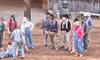A group of people in cowboy hats and work clothes gather around a person on the ground in front of a rustic barn.