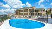 The image shows an outdoor swimming pool surrounded by lounge chairs, with a two-story building in the background under a partly cloudy sky.
