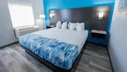 The image shows a modern hotel room with a large bed featuring a blue and white patterned comforter, set against a wood-paneled wall and accented by vibrant blue walls.