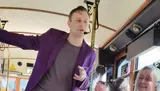 A man wearing a purple blazer and headset microphone is addressing an audience while holding onto a pole in what appears to be the interior of a bus or tram.