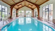 The image shows an indoor swimming pool with a vaulted ceiling, large windows, and several chairs along the poolside.