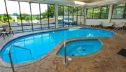 The image shows an indoor swimming pool and hot tub with a large windowed wall offering a view of the parking lot outside.