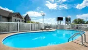 The image shows a clear blue swimming pool surrounded by a white fence, with a building and a sign visible in the background under a bright blue sky with scattered clouds.