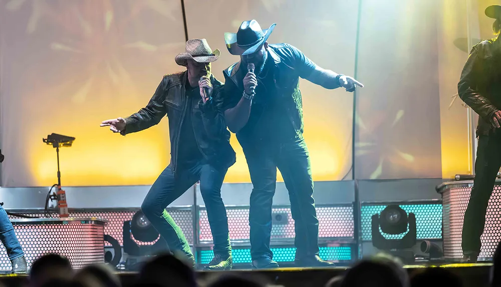 Two musicians in cowboy hats are energetically performing on stage with microphones in hand