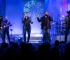 Four singers are energetically performing on stage with a band in the background illuminated by colorful stage lighting