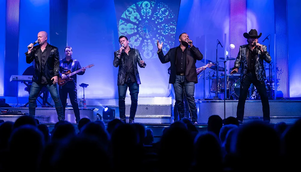 Four male vocalists are performing on stage with a live band and a vibrant light display in the background entertaining an audience