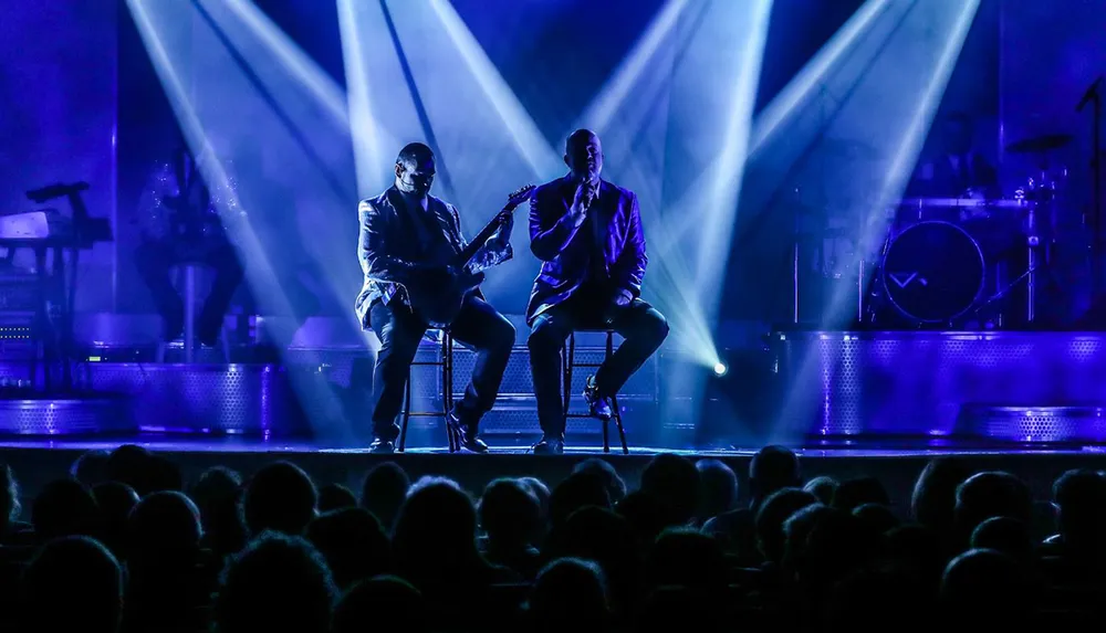 Two performers on stage are serenading an audience one playing guitar while the other sings with dramatic blue stage lighting creating a captivating atmosphere