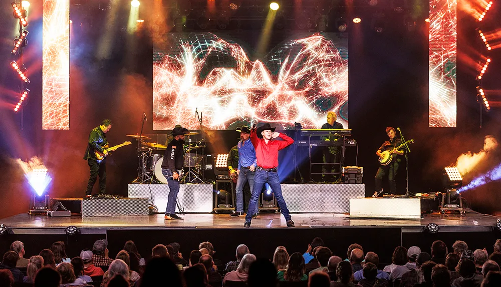 A country music band is performing energetically on stage with colorful lights and graphics in the background