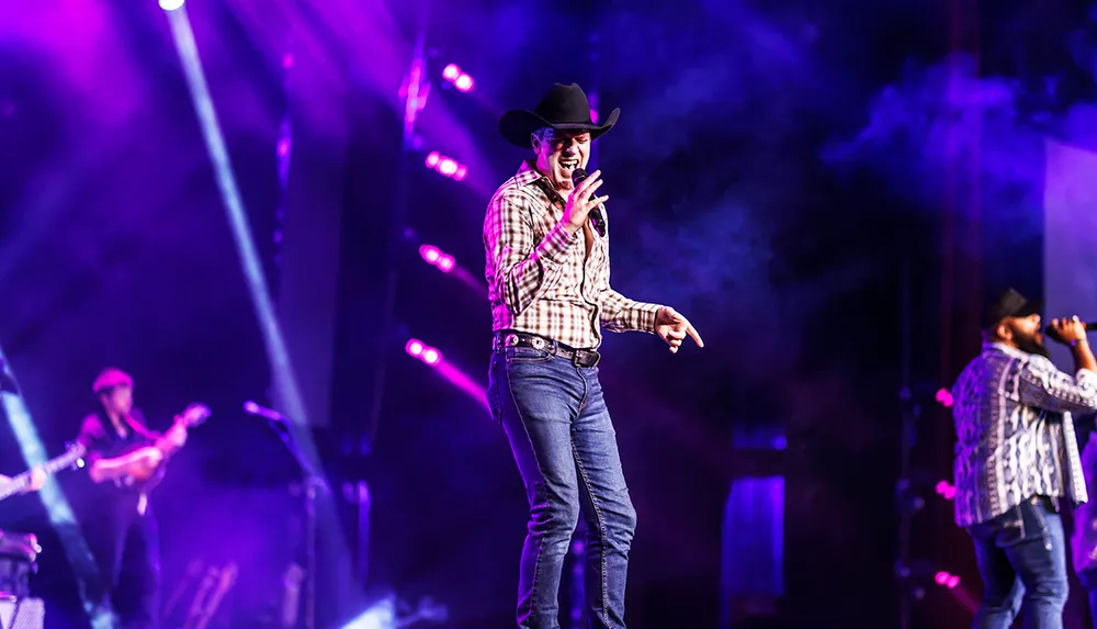 A performer in a cowboy hat energetically sings on a stage with colorful lights and smoke