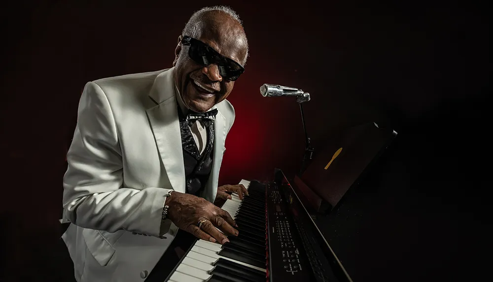 A joyful musician in a white suit plays the piano and smiles broadly behind sunglasses in a dimly lit setting