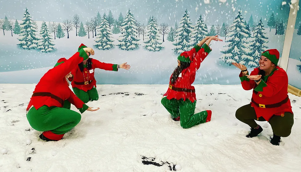 Three people dressed as elves are playfully throwing fake snow at each other in a room decorated to look like a winter wonderland