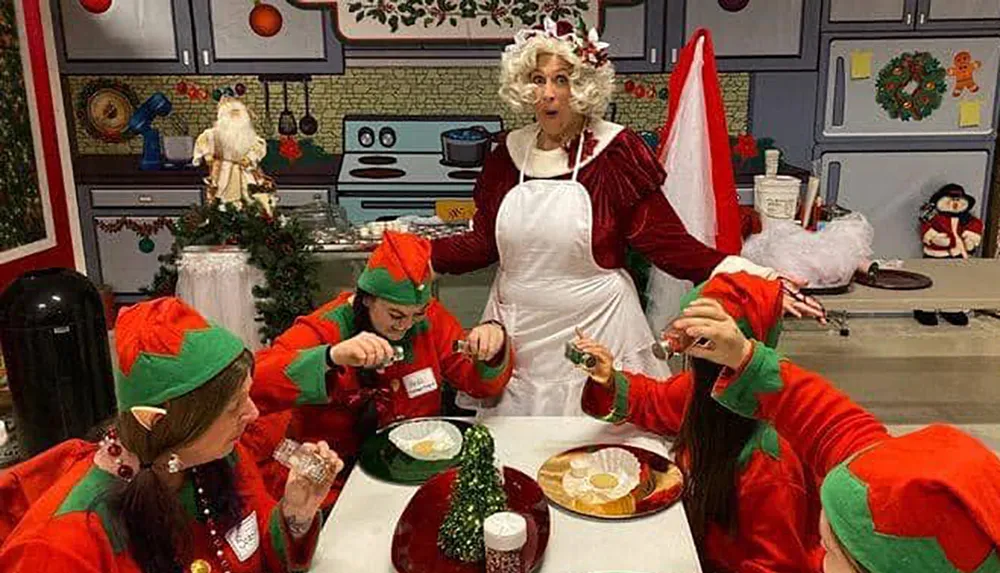 A person dressed as Mrs Claus looks surprised in a Christmas-themed kitchen while surrounded by individuals dressed as elves who are focused on preparing holiday treats