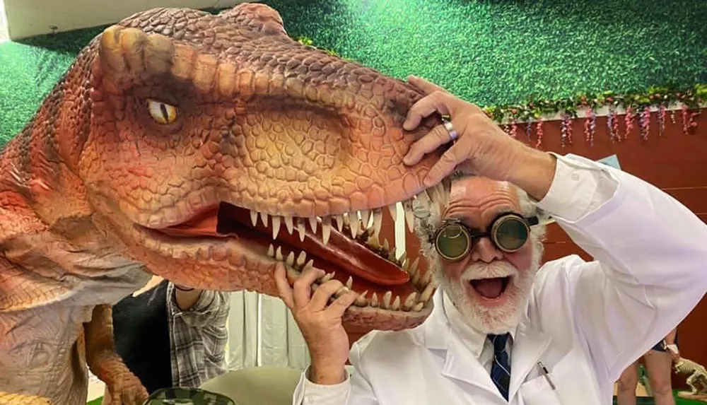 A person in a lab coat and goggles is playfully posing with a large model of a dinosaurs head