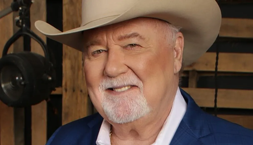 The image shows a smiling man with a white mustache and beard wearing a cowboy hat and a blue blazer with a rustic wooden background