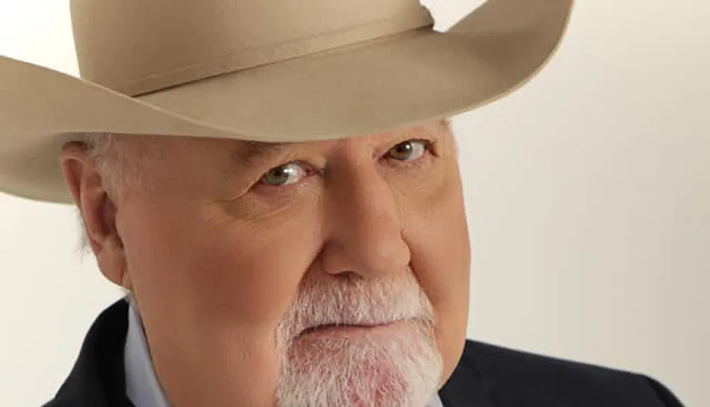 The image shows a close-up of an older man with a grey beard wearing a large beige cowboy hat and a black jacket set against a cream background