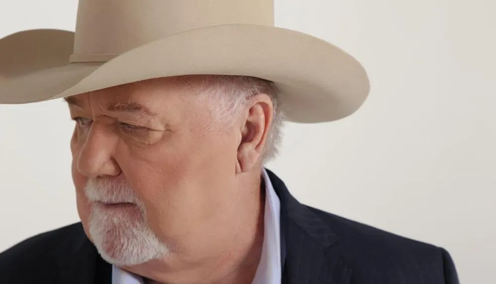 A man with a mustache wearing a beige cowboy hat looks to the side with a serious expression against a light background