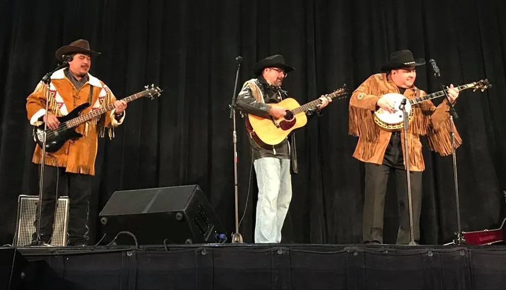 Three musicians dressed in cowboy hats and fringed jackets are performing on stage with a banjo an acoustic guitar and a bass guitar