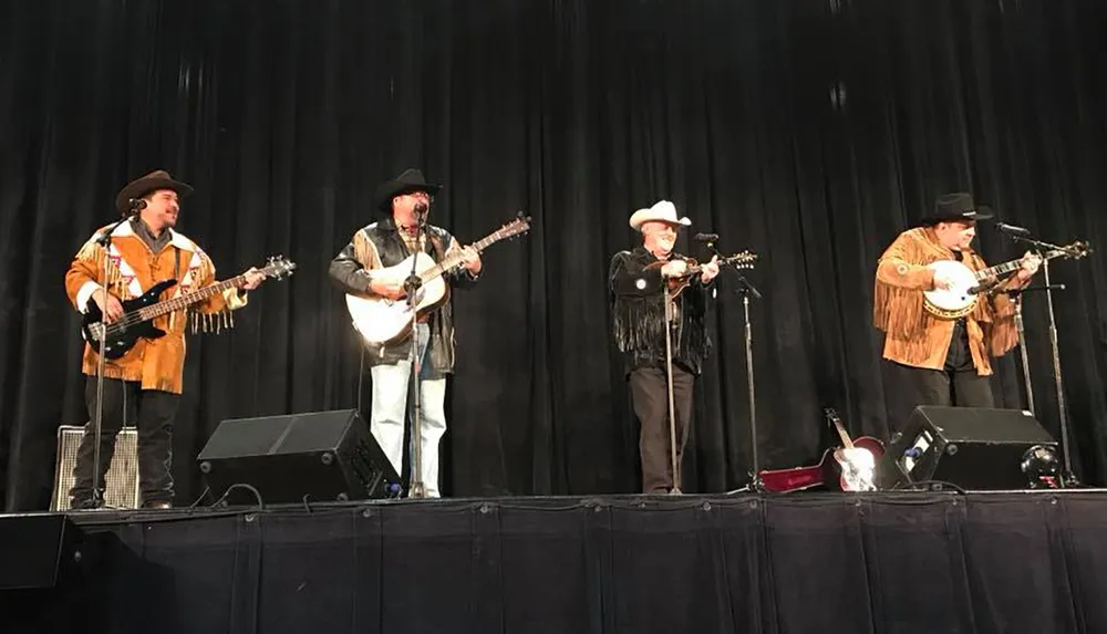 A band of four musicians each wearing cowboy hats and Western attire is performing on stage with a black backdrop playing instruments that include a banjo and guitars