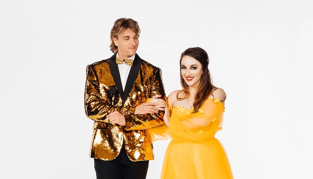 A man in a sparkling gold sequin jacket and a woman in a flowing yellow dress are smiling and posing together against a white background