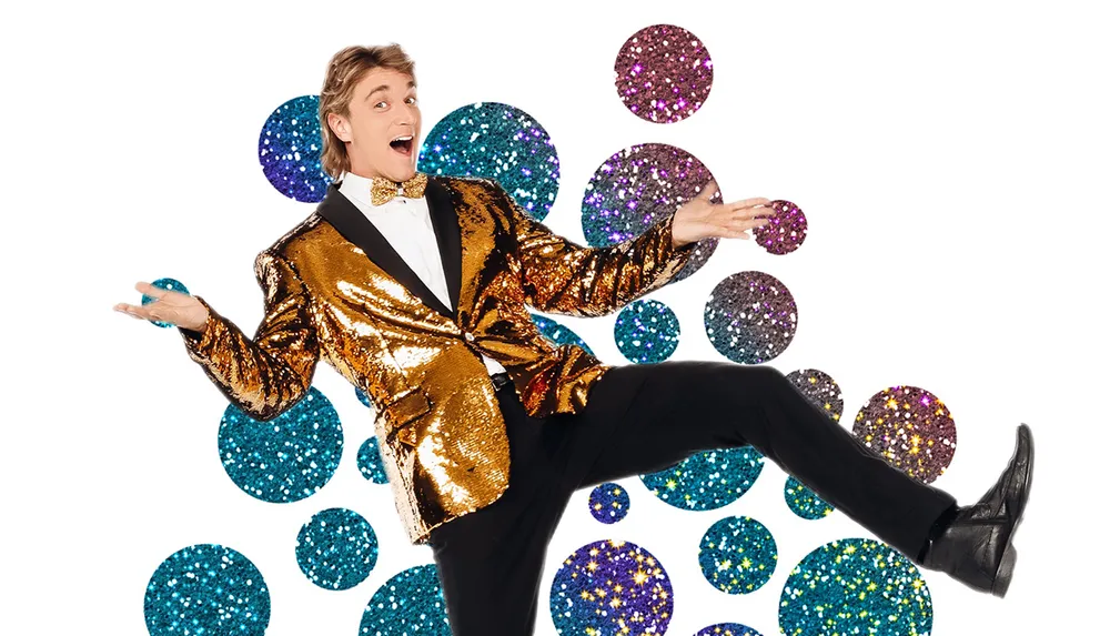 A person in a glittering gold jacket and black pants is energetically posing with an excited expression against a background of colorful sparkling dots