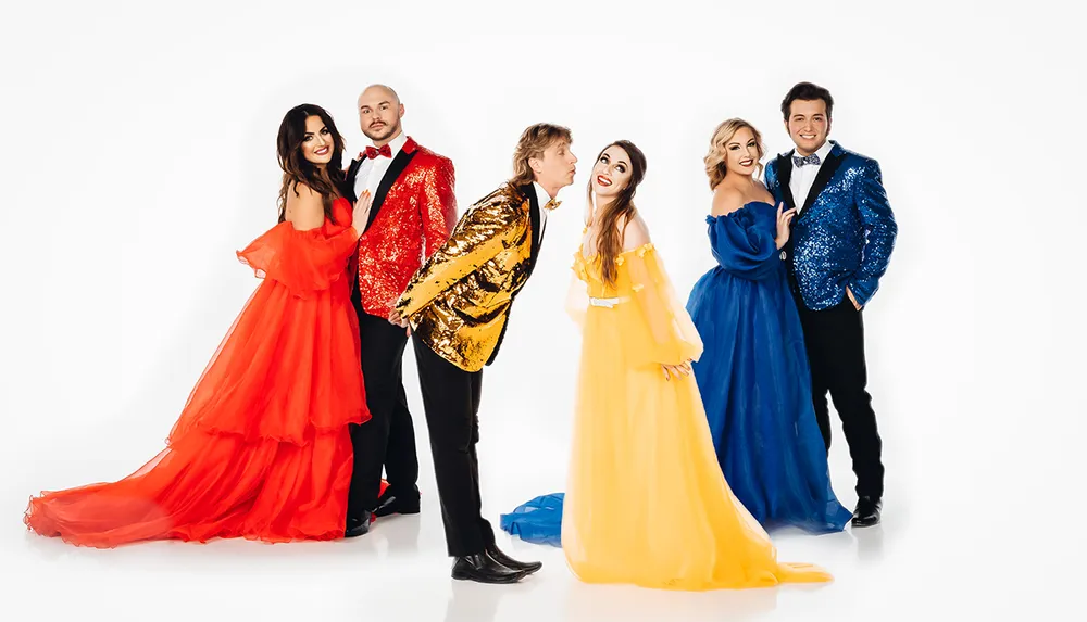 Five people wearing colorful formal attire pose in front of a white background with two couples on the sides and a man in the center playfully pouting towards a woman in yellow