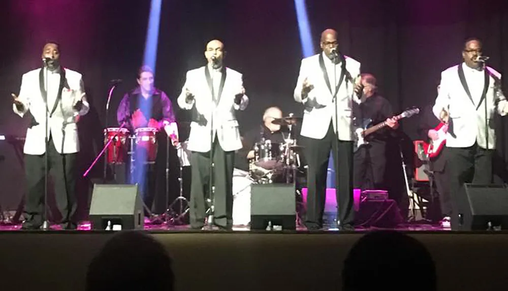 Four singers in white jackets are performing onstage with a band that includes a drummer guitarist and percussionist under colored stage lighting