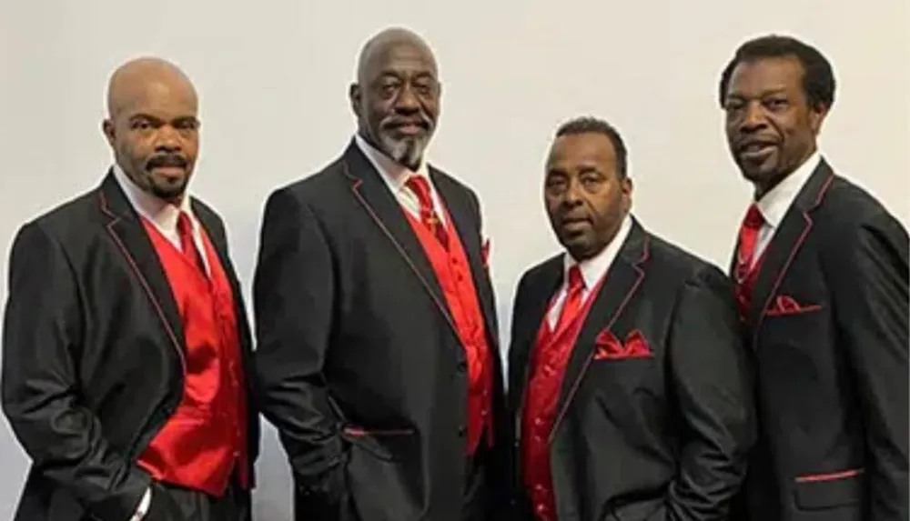 Four men dressed in matching black suits with red shirts and red pocket squares are standing together posing for a photo
