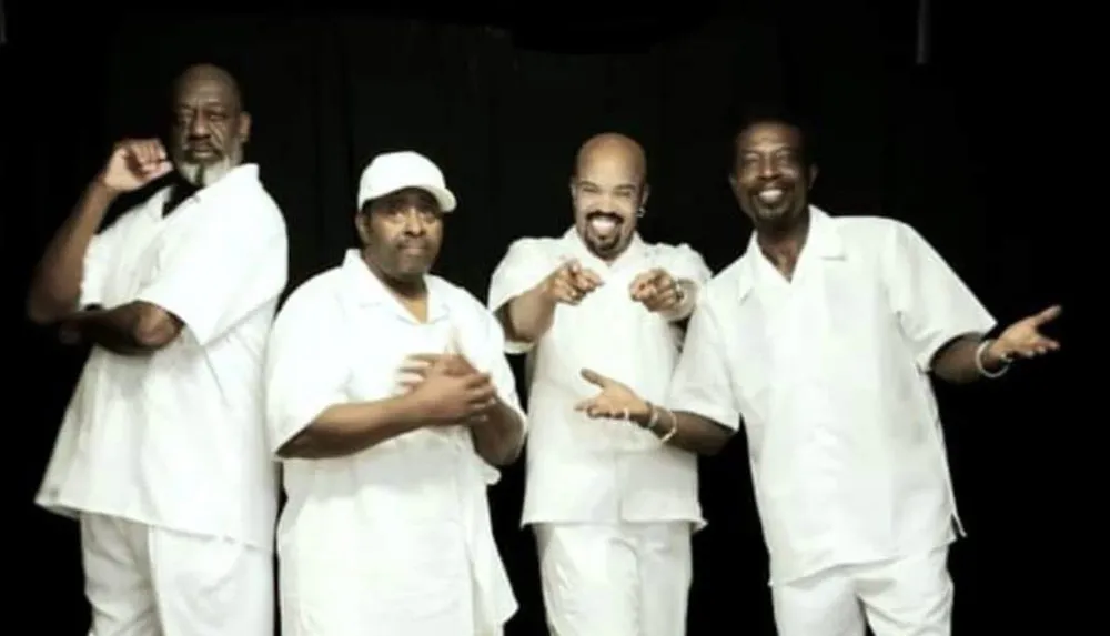 Four smiling men dressed in white are playfully posing against a black background
