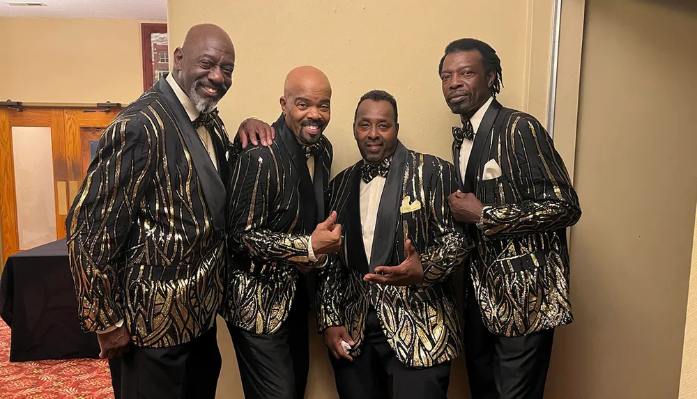 Four smiling men in matching black and gold patterned suits pose for a group photo