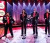 Four men in sparkly suits and red shirts are performing on stage under bright lights each holding a microphone