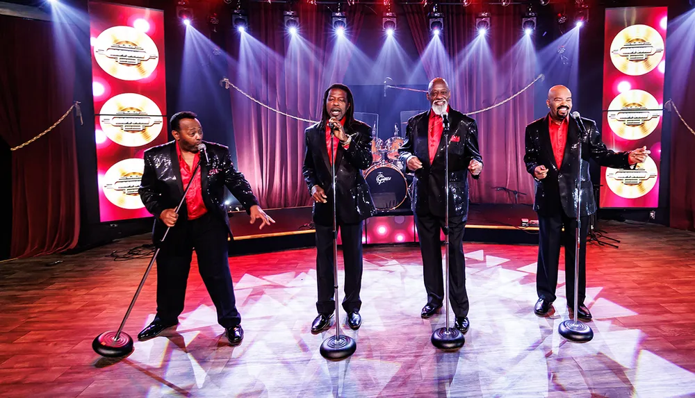 Four men in shiny jackets perform energetically on a vibrant illuminated stage
