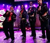Four men in sparkly suits and red shirts are performing on stage under bright lights each holding a microphone