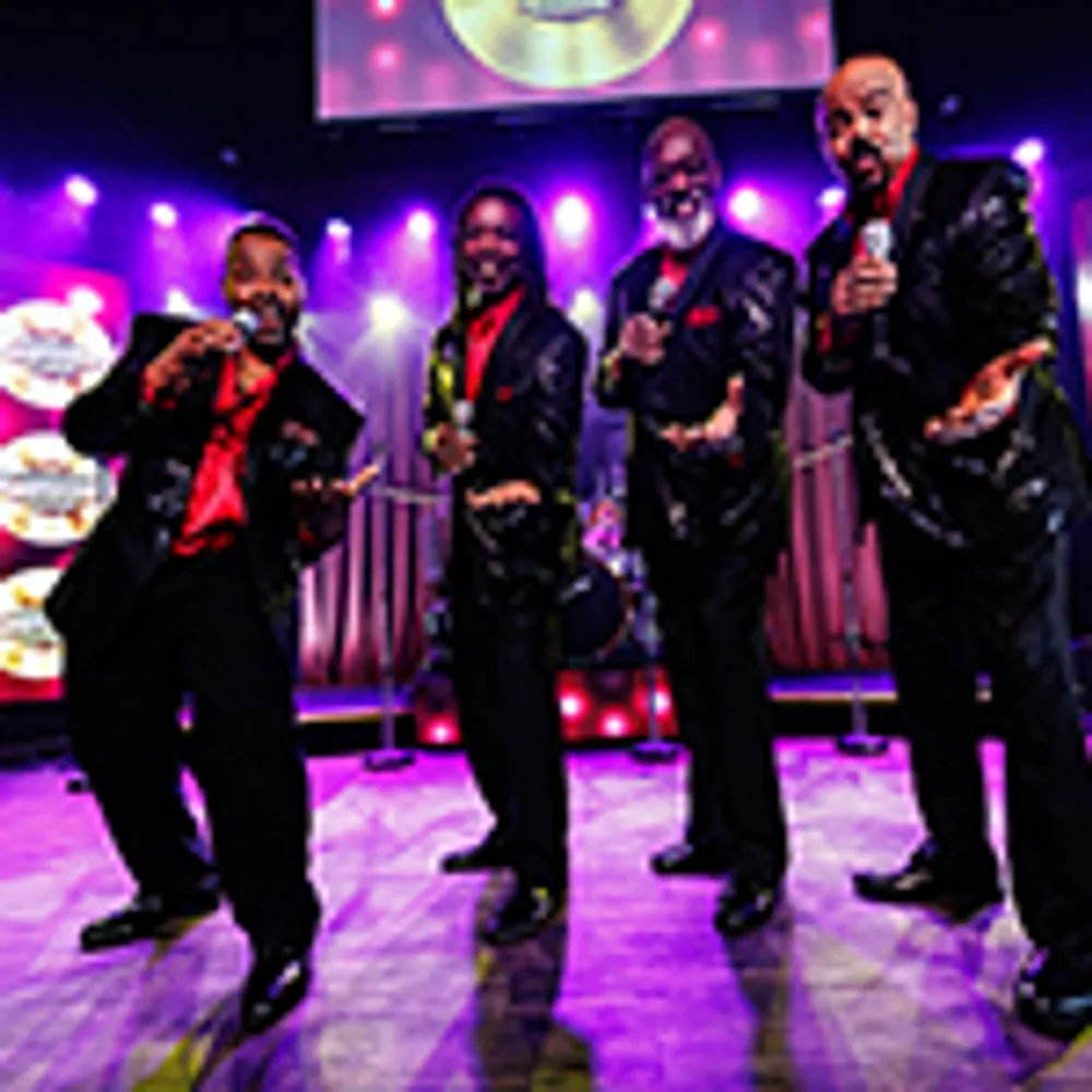 Four performers dressed in black suits and red shirts enthusiastically pose on stage with purple lighting and a backdrop featuring a logo