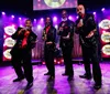 Four men in sparkly suits and red shirts are performing on stage under bright lights each holding a microphone