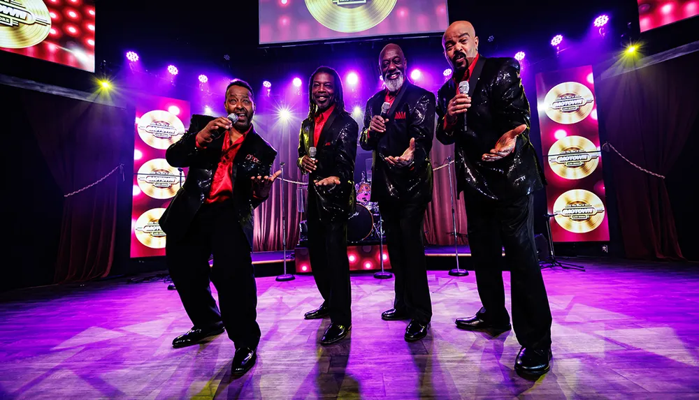 Four men in sparkly suits and red shirts are performing on stage under bright lights each holding a microphone