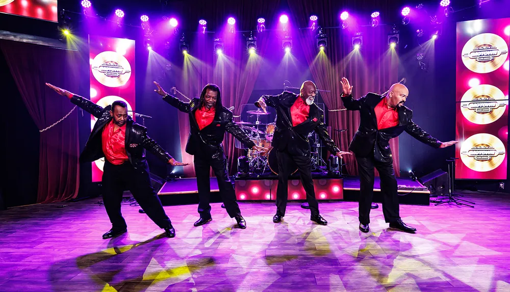 Four performers in black suits with red shirts energetically dance on a stage lit with purple lights under a backdrop displaying Motown records