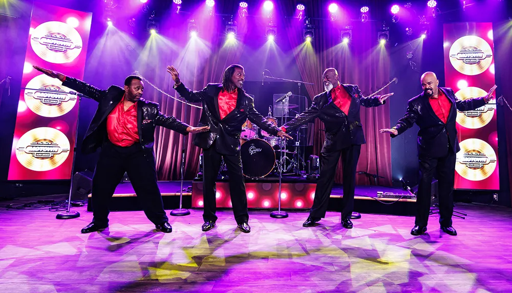 Four performers in matching black suits with red shirts energetically dance on a stage with bright purple lighting