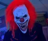 A person is wearing a scary clown mask with bright red hair and a wide menacing grin standing indoors under a blue light
