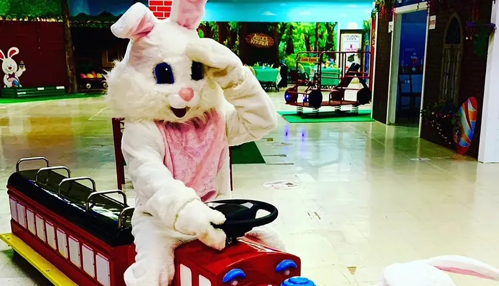 A person dressed in a large fluffy bunny costume is seated on a red childrens ride-on train in an indoor play area