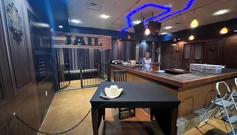 The image depicts an interior space styled with a Western theme featuring a JAIL sign above bars and a table set for dining in the foreground with a person standing behind a counter possibly set up as a bar or serving area in the background