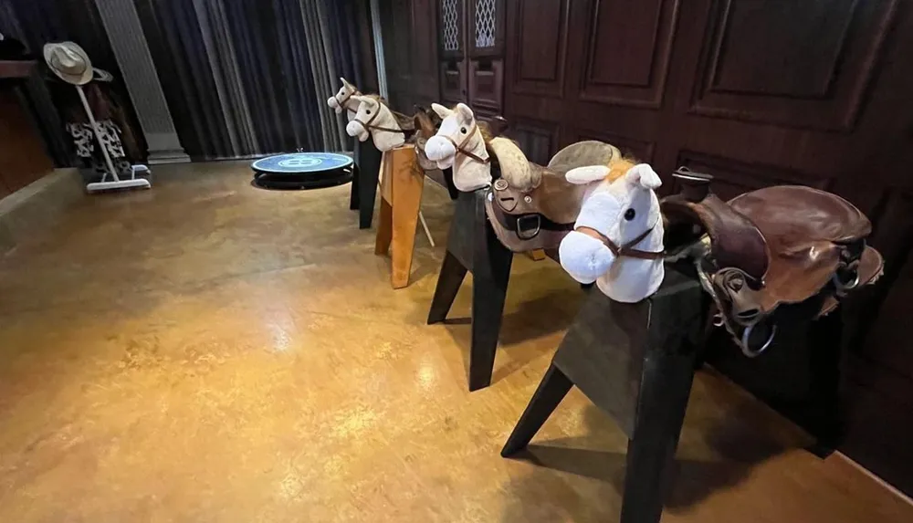 A row of plush hobby horses are arranged in a room each sporting a different colored saddle and bridle with a decorative hat stand visible in the background