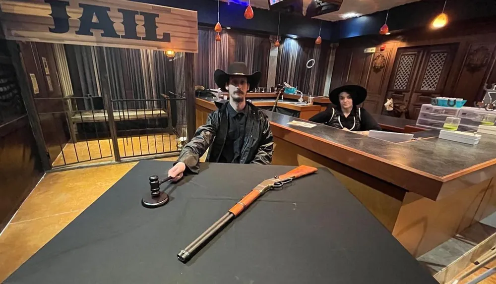Two individuals are dressed in western attire facing the camera with one seated behind a counter and the other standing with a gavel in hand in a room themed to look like a Wild West courtroom with a rifle on the table and jail cells in the background