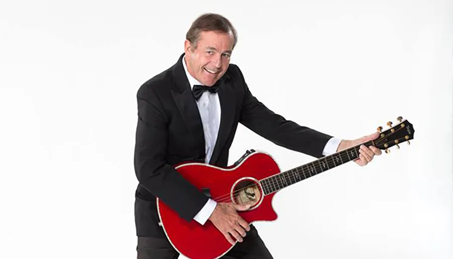 A man in a black suit and bow tie is playfully posing with a red guitar against a white background.