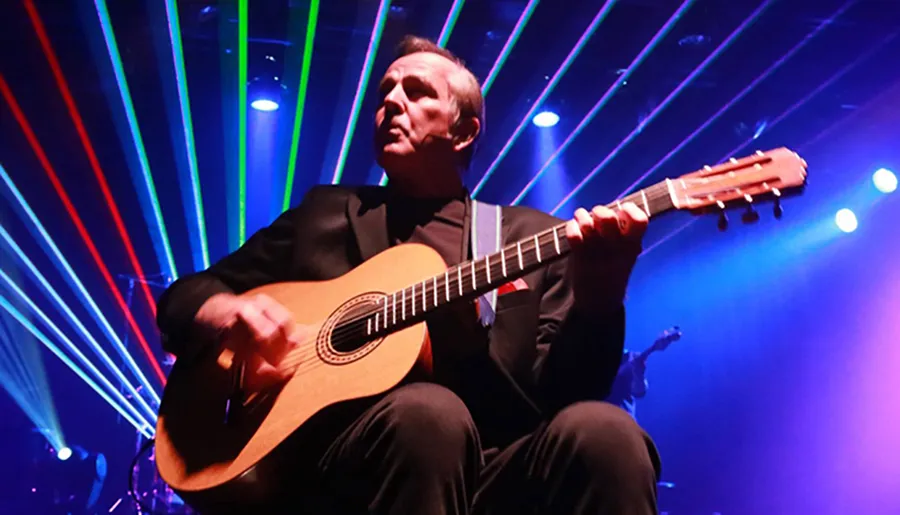 A musician in a suit passionately plays an acoustic guitar on stage amid a vibrant backdrop of colorful laser lights.