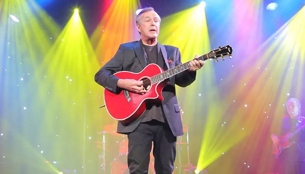 A musician performs onstage with a red acoustic guitar under a vibrant display of stage lights