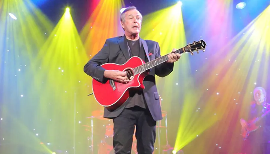A musician performs onstage with a red acoustic guitar under a vibrant display of stage lights.