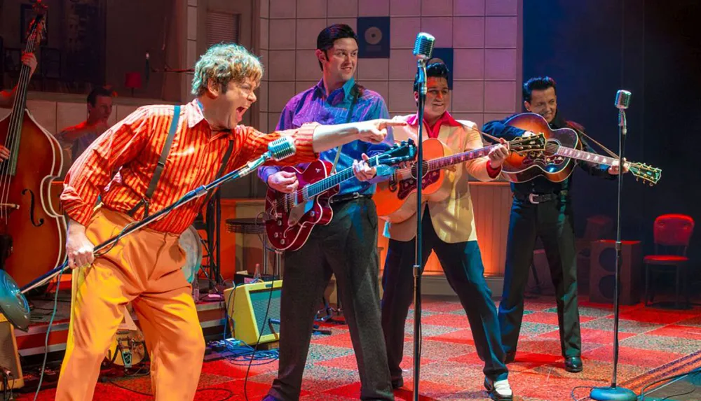 Four performers in a vintage setting energetically play guitars and a double bass on stage evoking the style of a 1950s or 1960s rock-and-roll band